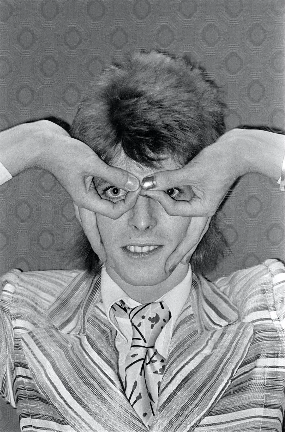 David Bowie in a striped jacket and patterned tie, grinning, with his hands turned upside down over his face: forefingers and thumbs curled into rings around his eyes, the rest of his fingers resting on his cheeks.