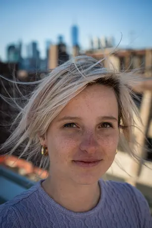 A woman with windblown platinum hair on a rooftop, the Manhattan skyline soft and blue behind her.