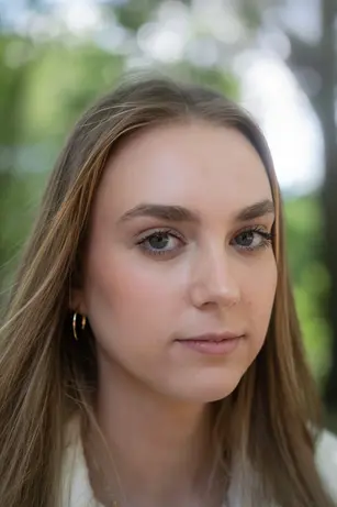 A close portrait of a young woman with long fair hair and a gold hoop earring, looking just past the camera with a level, unsmiling expression, summer foliage blurred to green behind her.