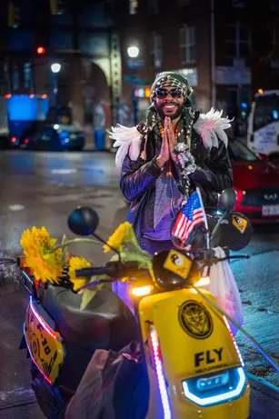 A man in a patterned headwrap, sunglasses and feathered wings presses his palms together in greeting behind a yellow scooter strung with sunflowers and LED lights on a wet night street.