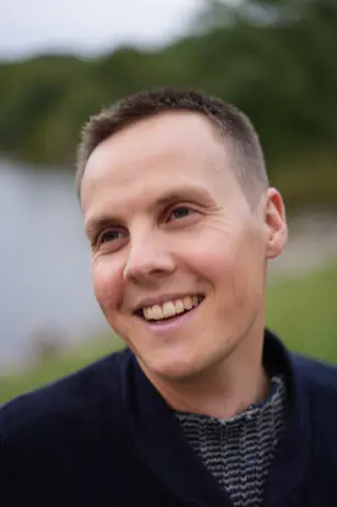 A man in a navy jacket smiles off to one side, a lake and dark trees soft in the background.