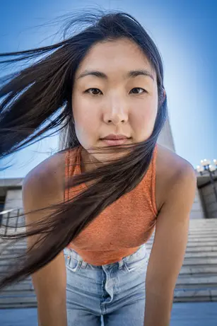 Shot from below, a woman leans down toward the camera on a flight of pale stone steps, her long dark hair blown sideways across her face, in an orange cropped top against a clear blue sky.