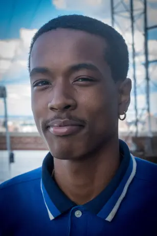 A young man in a blue polo shirt with a gold hoop earring tilts his head and smiles slightly, a painted sky of clouds and rooftop scaffolding behind him.