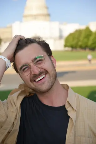A man in round gold glasses laughs with his eyes shut and a hand on his head, a streak of green paint on one cheek and an event wristband on his wrist, a park and a domed building behind him.
