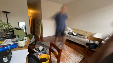 A timelapse of two people assembling a wooden sauna in a living room, from flat panels leaning against the wall to a finished cabin beside the sofa.